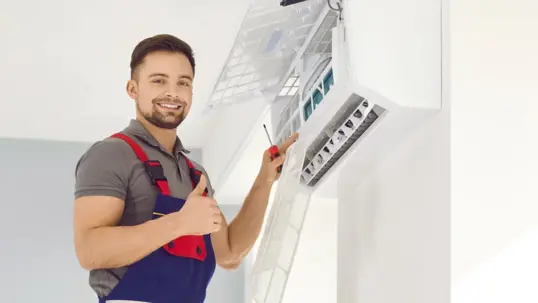 A smiling technician works on an AC unit, giving a thumbs-up
